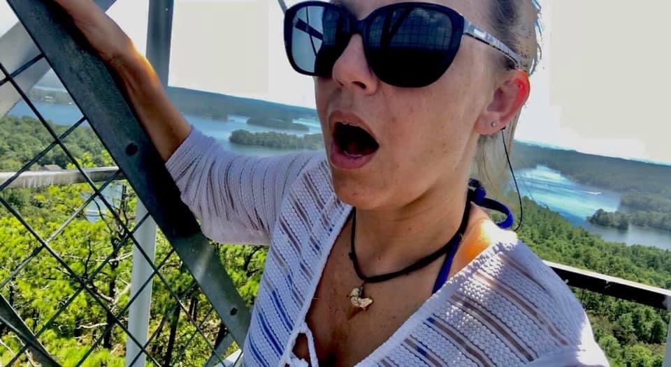 Close up photo of Shaylee from Pelican Point Expeditions in sunglasses with her mouth dropped. She is climbing the steps of the Smith Mountain Fire Tower. Trees and a lake are in the background