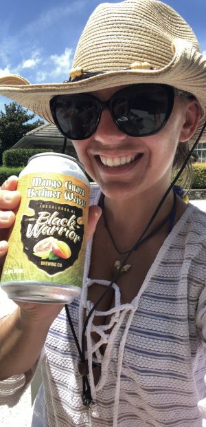 Shaylee from Pelican Point Expeditions poses and smiles in sunglasses and a cowboy hat while holding up a can of Black Warrior Brewing Company Mango Guava